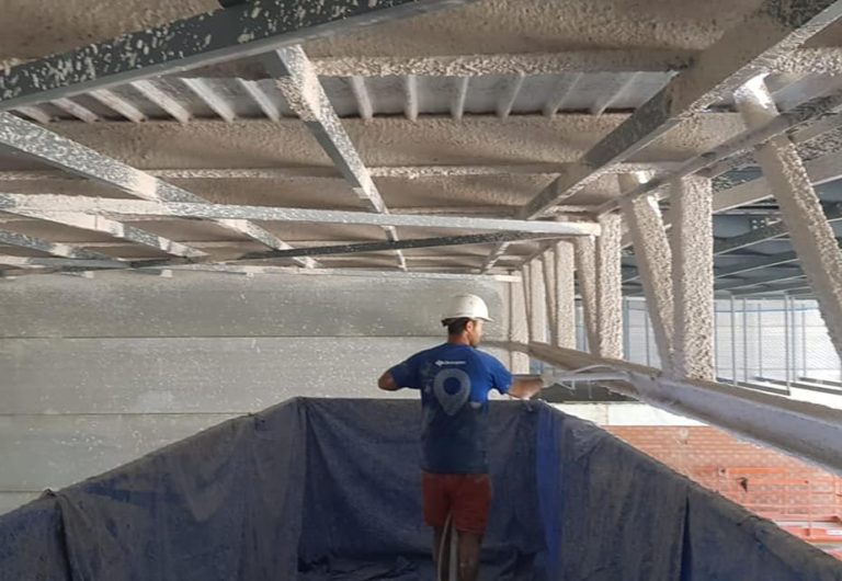 Un trabajador de la construcción, con casco blanco y camisa azul, de pie sobre una plataforma, rocía material aislante o MORTERO sobre vigas metálicas y el techo en el interior de un edificio inacabado.