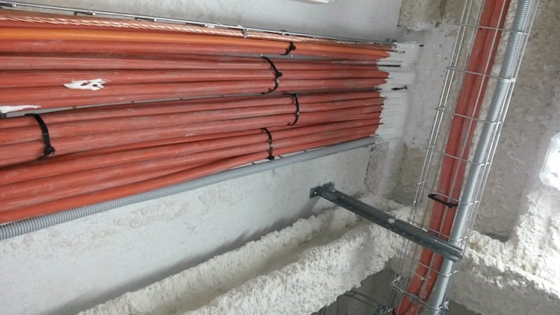 Varios conductos eléctricos de color naranja, con sellados resistentes al fuego, están perfectamente atados y asegurados con bridas negras, recorriendo una pared y un techo con aislamiento de espuma pulverizada. Una bandeja metálica sujeta verticalmente algunos de los conductos.